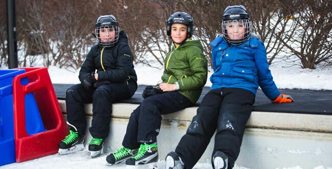 Three boys sitting on the side of an outdoor skating rink in winter